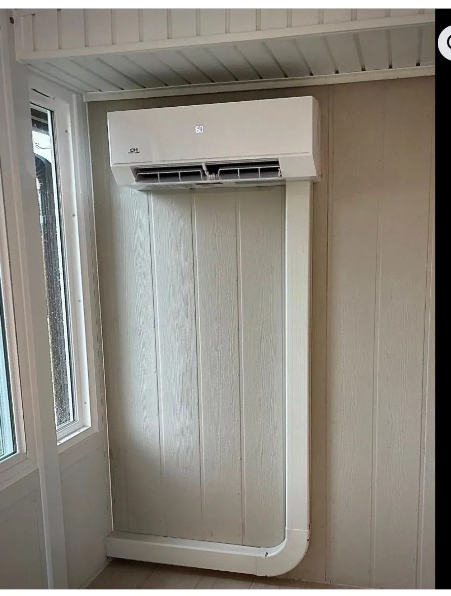 White wall-mounted air conditioner above a radiator in a beige enclosed porch by a window.