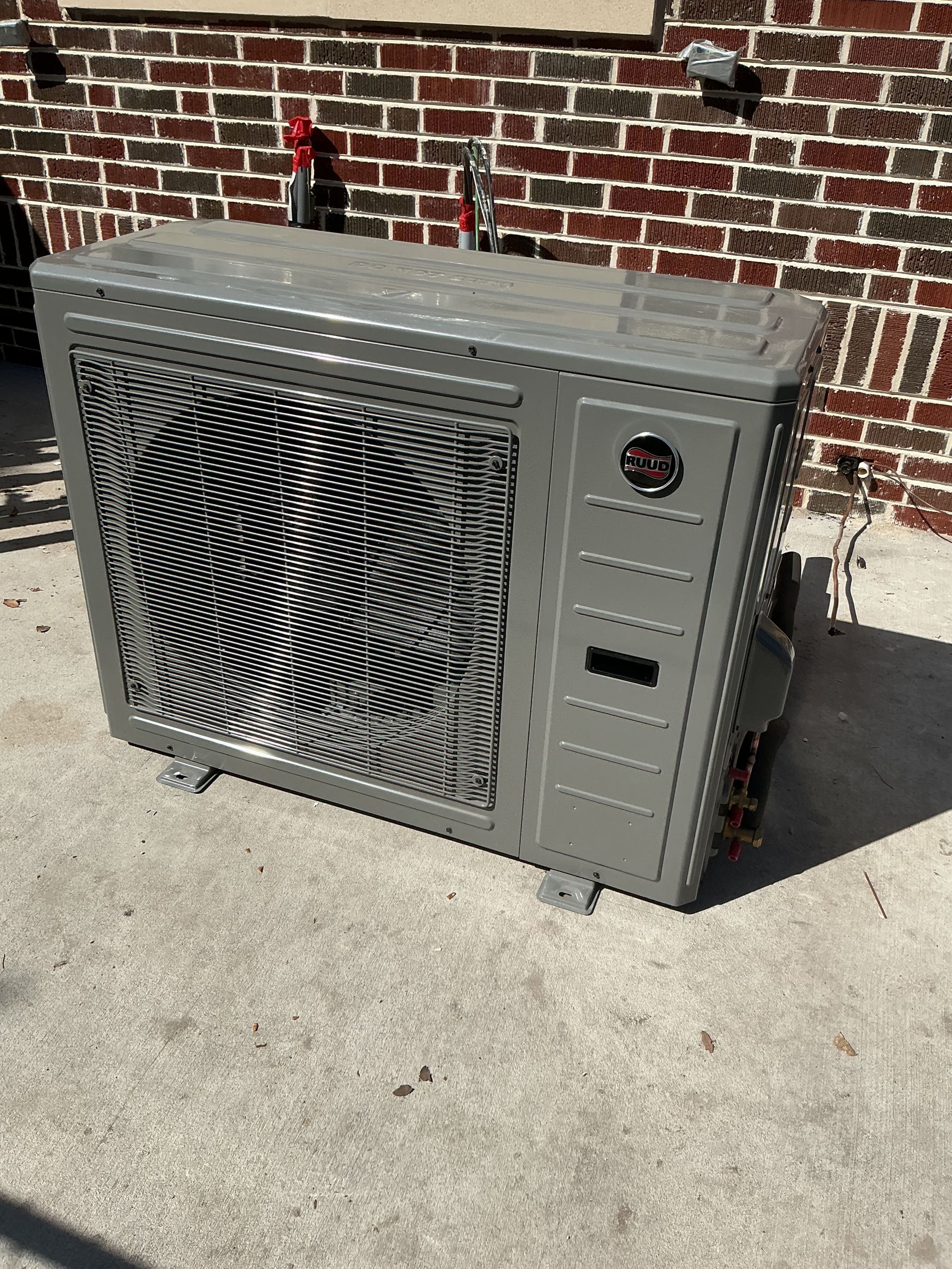 Gray outdoor HVAC unit beside a brick wall on a concrete patio, with a mesh front panel and control dials.