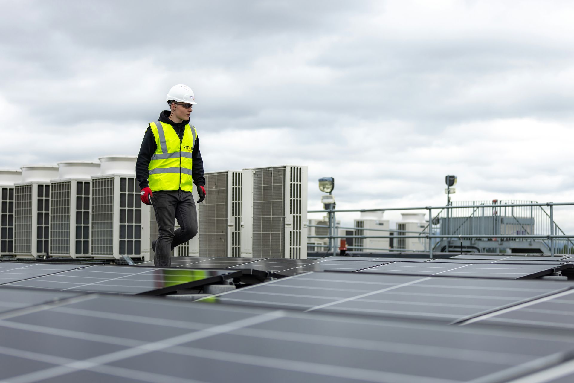 Worker in a hard hat and high-vis vest walking on a rooftop solar panel array under cloudy skies