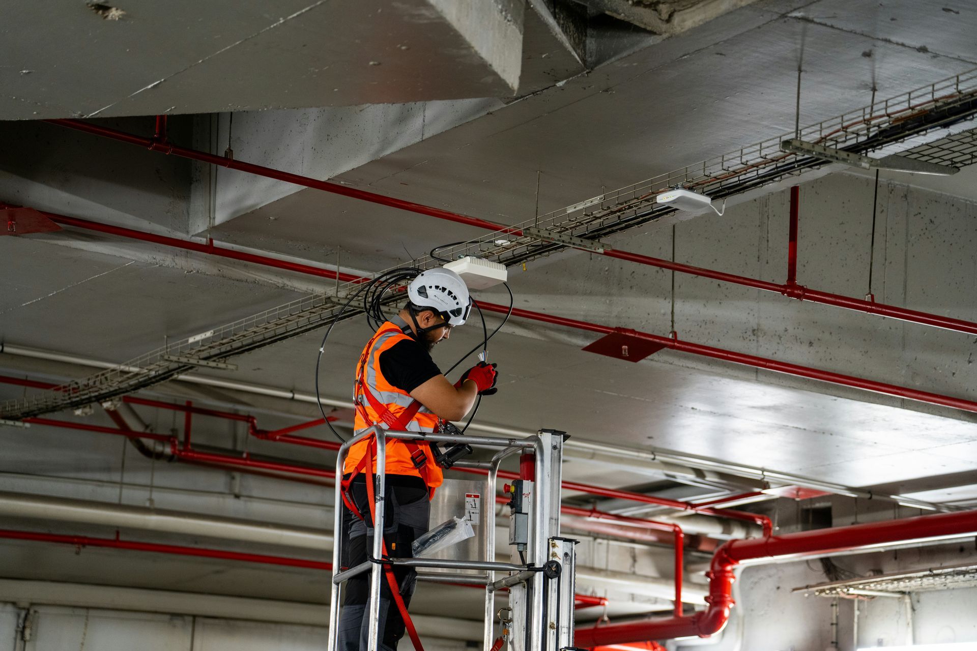 Worker in orange safety vest inspecting ceiling pipes on a scaffold in a parking garage