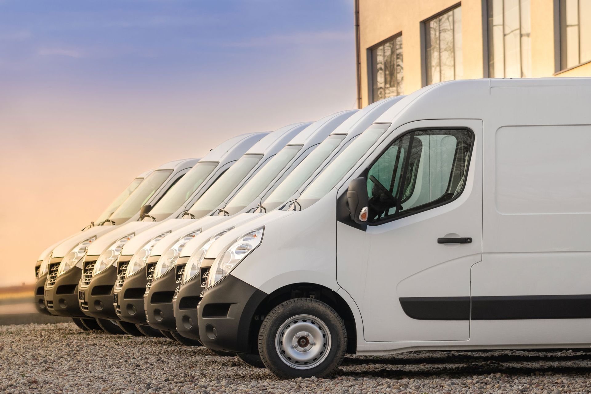 Row of white cargo vans parked outside a building, against a sunset sky.