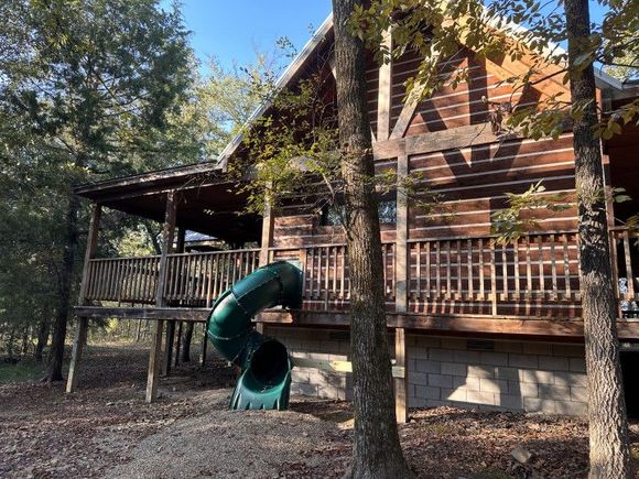 Wooden cabin with porch and green slide in a wooded yard, sunlight filtering through trees