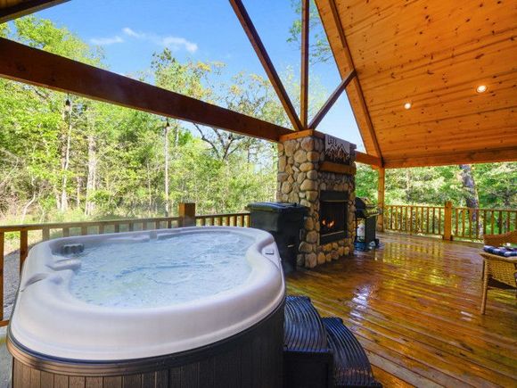 Covered wooden deck with a hot tub, stone fireplace, and forest view