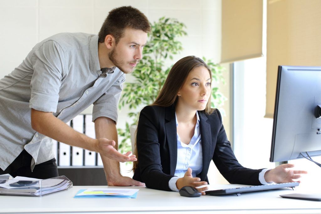 Two coworkers review documents at a desk beside a desktop computer in a bright office