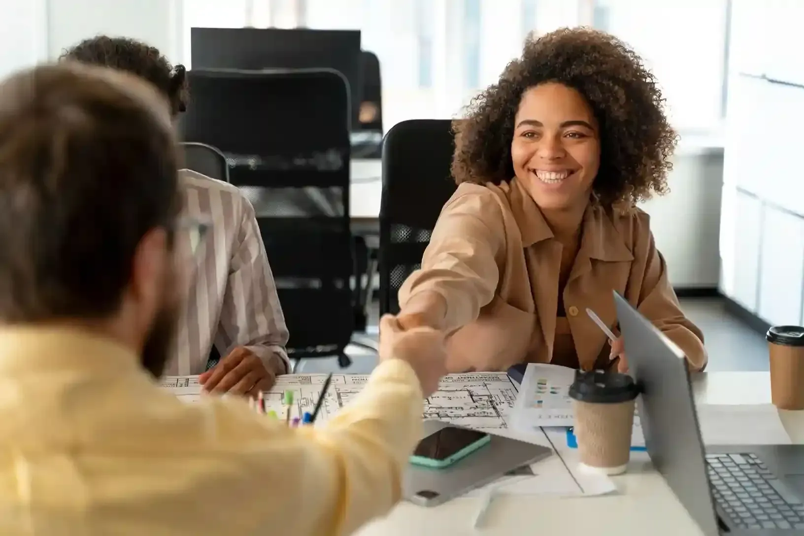 Colleagues in a bright office shaking hands across a desk, with papers, coffee cups, and a laptop nearby
