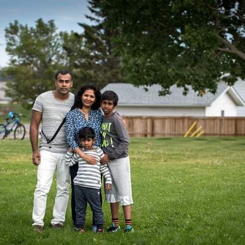Family of four posing on a grassy field with trees and houses in the background.