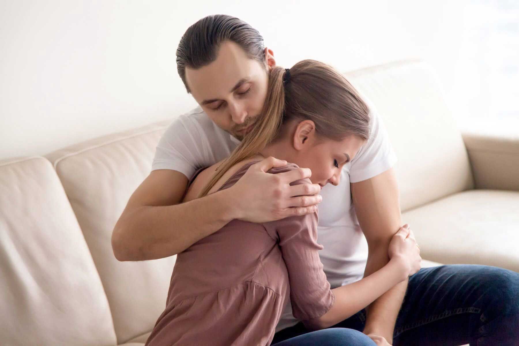 Two people sit on a couch, embracing with comforting expressions in a bright living room.