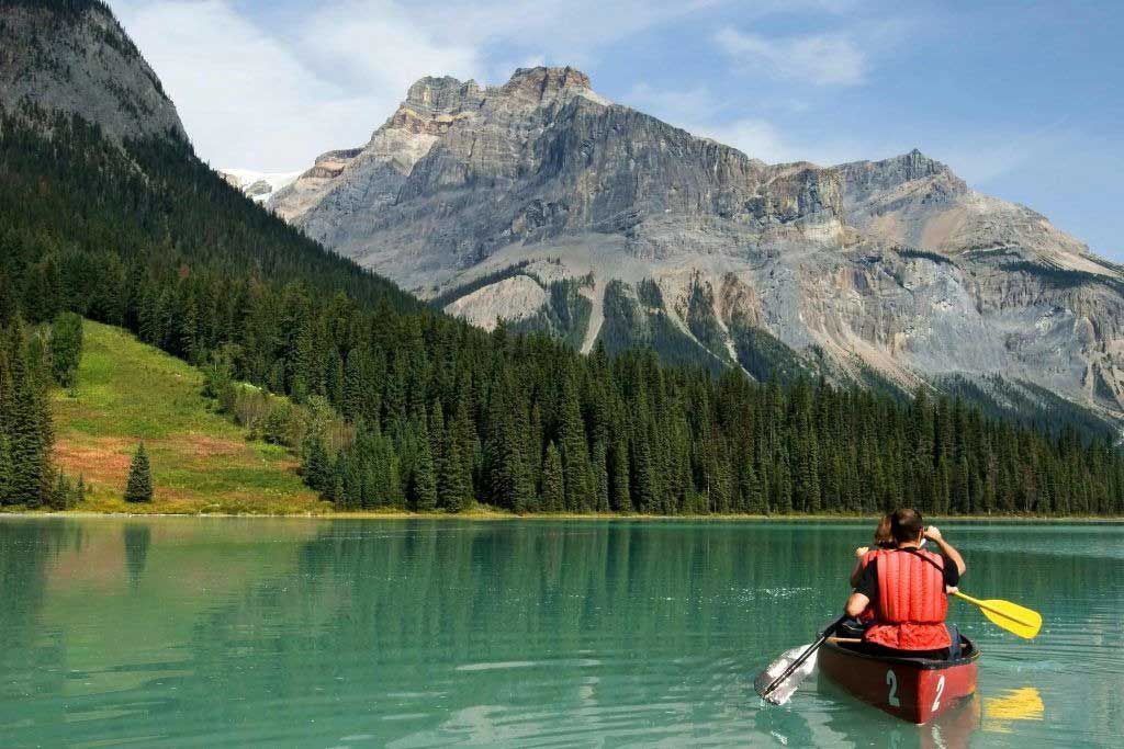 Kayaker in an orange life vest paddles a turquoise lake with forested mountains and cliffs behind.