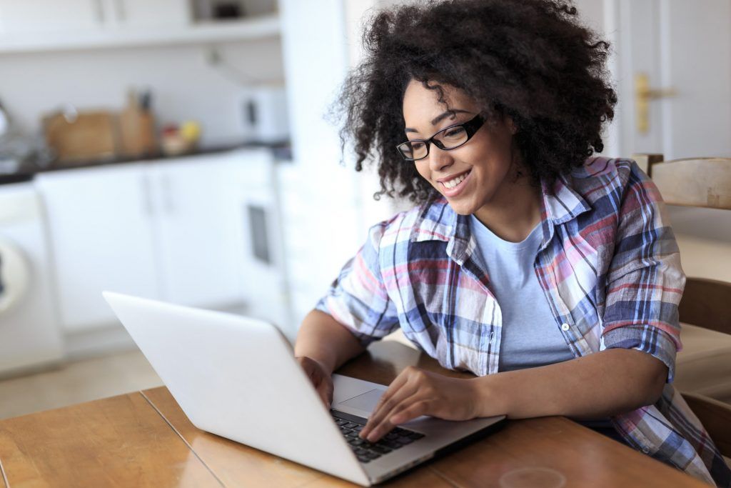 Person smiling and using a laptop at a wooden table in a bright kitchen