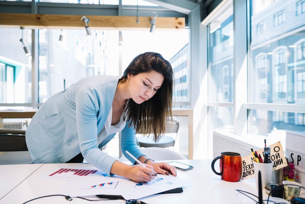 Woman leaning over a desk, writing on papers in a bright office with mugs and art supplies nearby