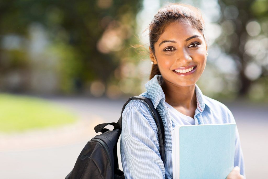 Smiling student with backpack holding a notebook outdoors on a sunny campus path