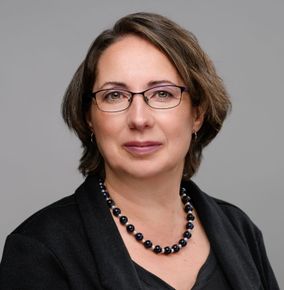 Studio headshot of a woman wearing glasses, a dark blazer, and a beaded necklace on a gray background