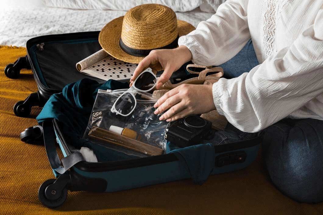 Hands packing clothes into an open suitcase beside a straw hat on a bed
