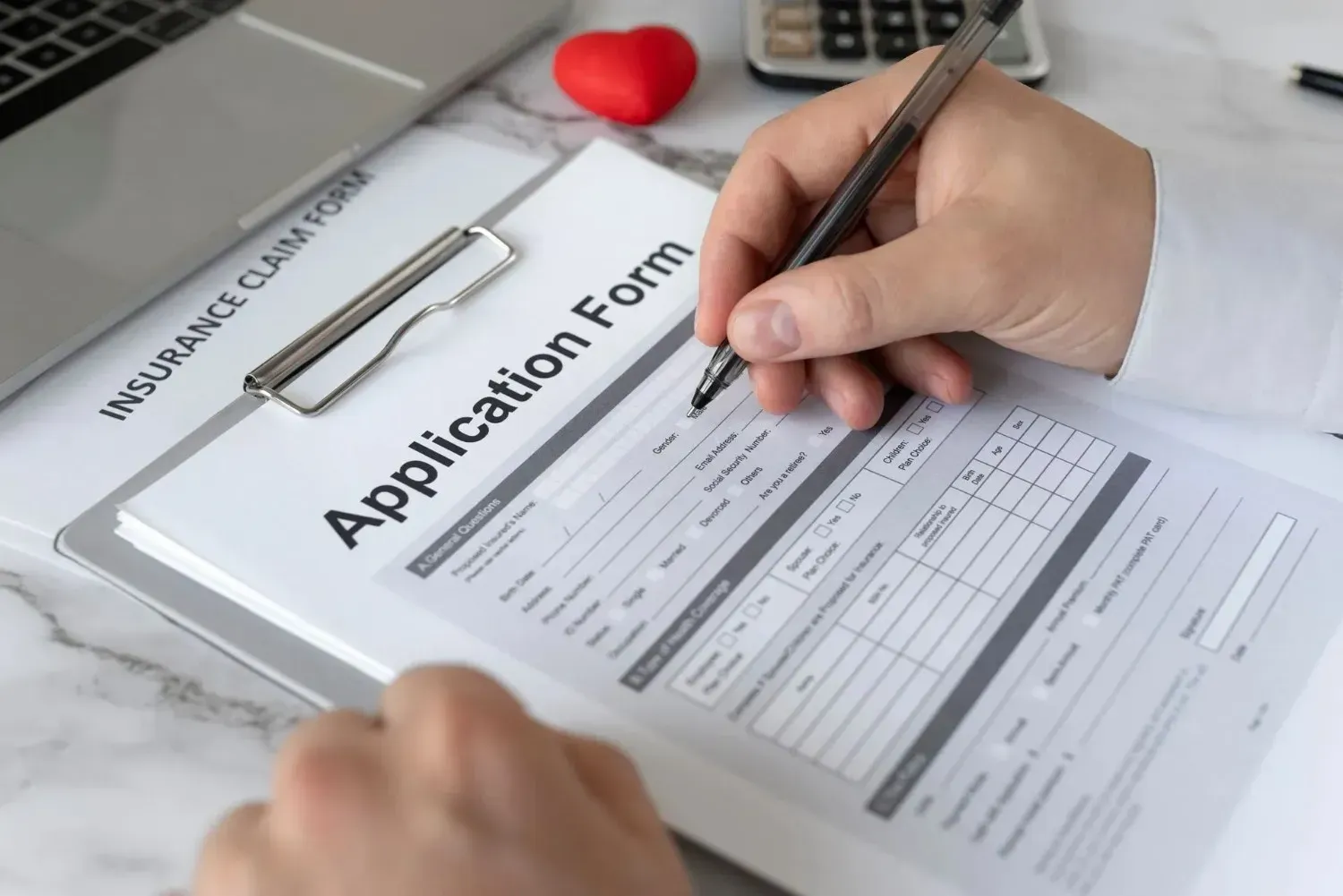 Hands filling out an application form on a clipboard with a pen next to a calculator and red heart