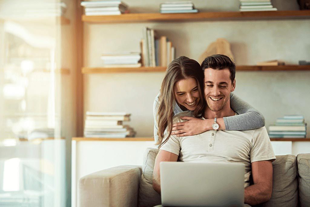 Couple smiling on a couch, looking at a laptop in a bright living room