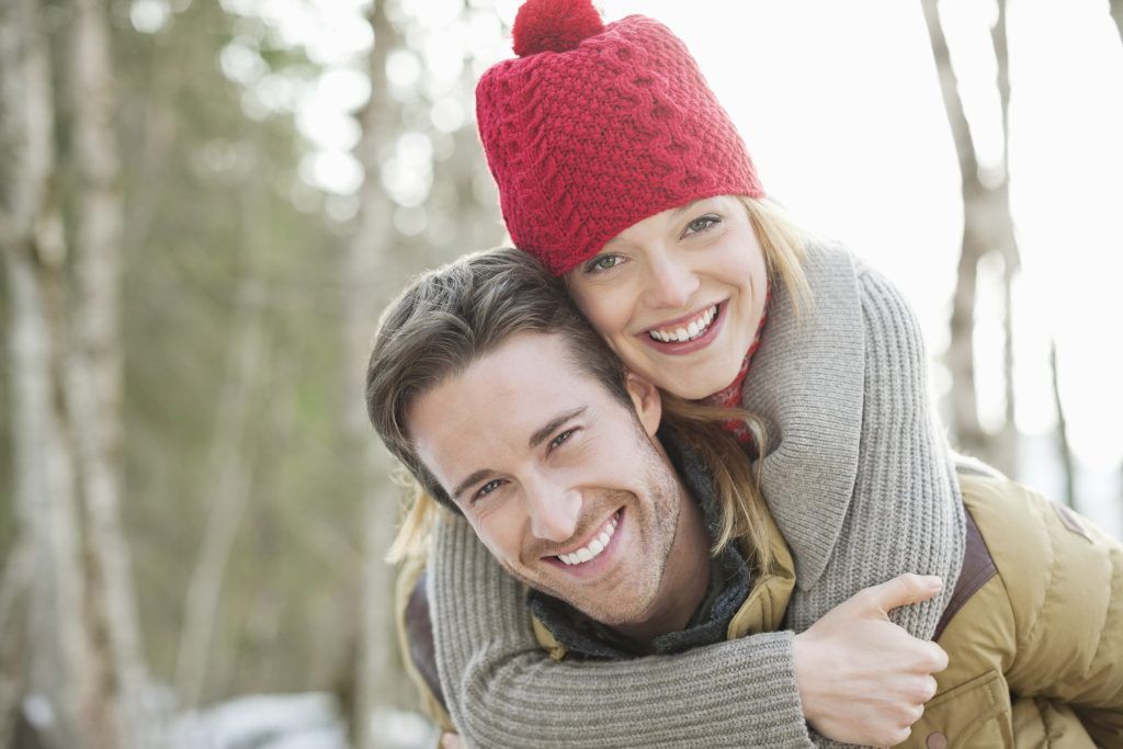 Smiling couple outdoors in winter, woman in a red hat hugging a man from behind.