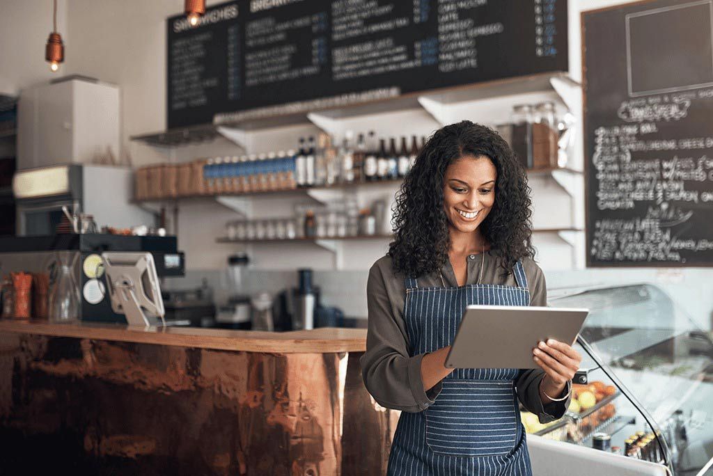 Woman in an apron using a tablet behind a cafe counter with menu boards in the background
