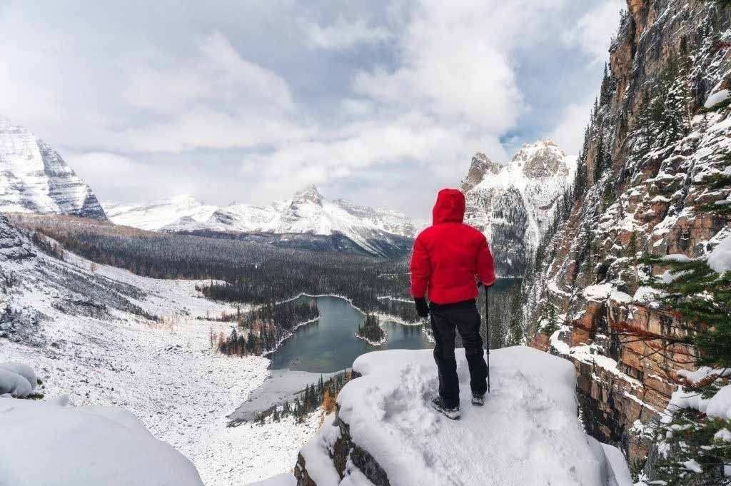 Person in a red jacket standing on a snowy cliff overlooking a mountain lake and valley