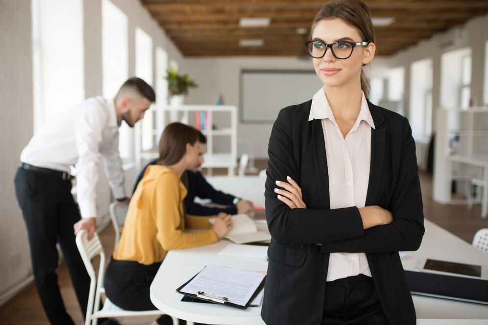 Confident professional in black suit standing with arms crossed in a modern office while coworkers work behind her