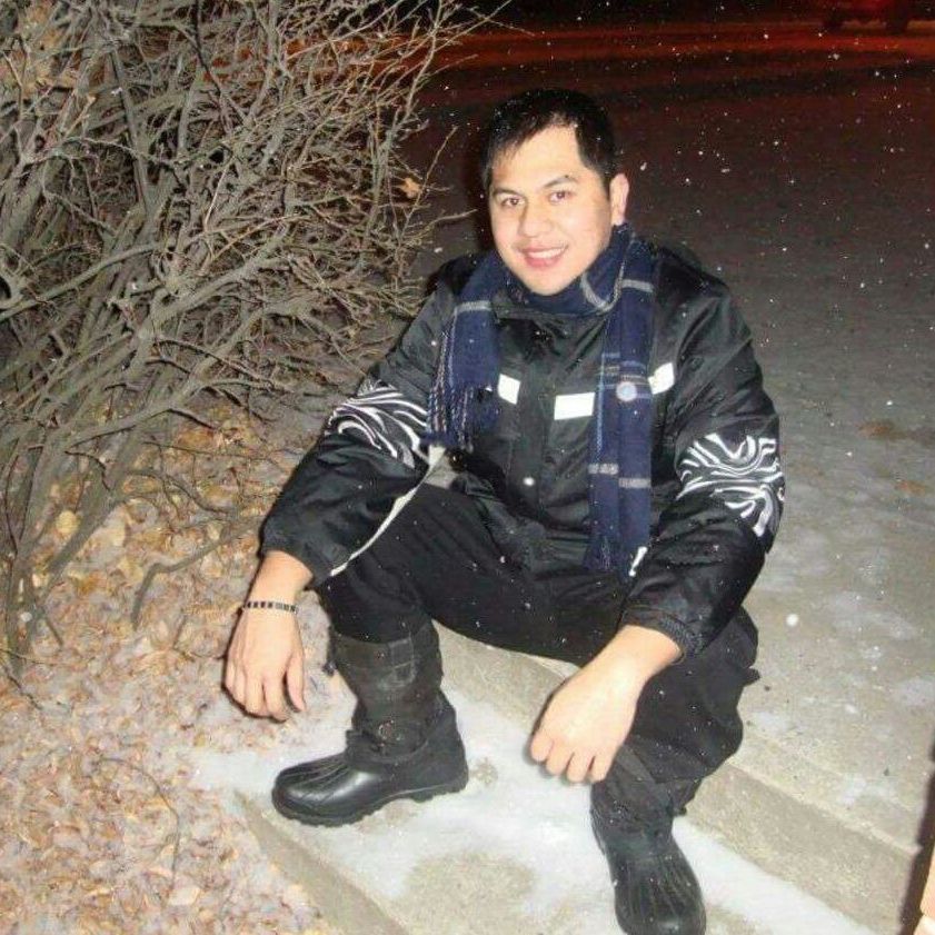 Person crouching in the snow at night, wearing a black jacket and boots, smiling near a bush