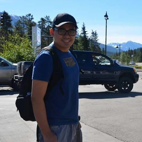 Smiling person in blue shirt and cap with backpack standing on a sunny street near parked trucks and mountains.