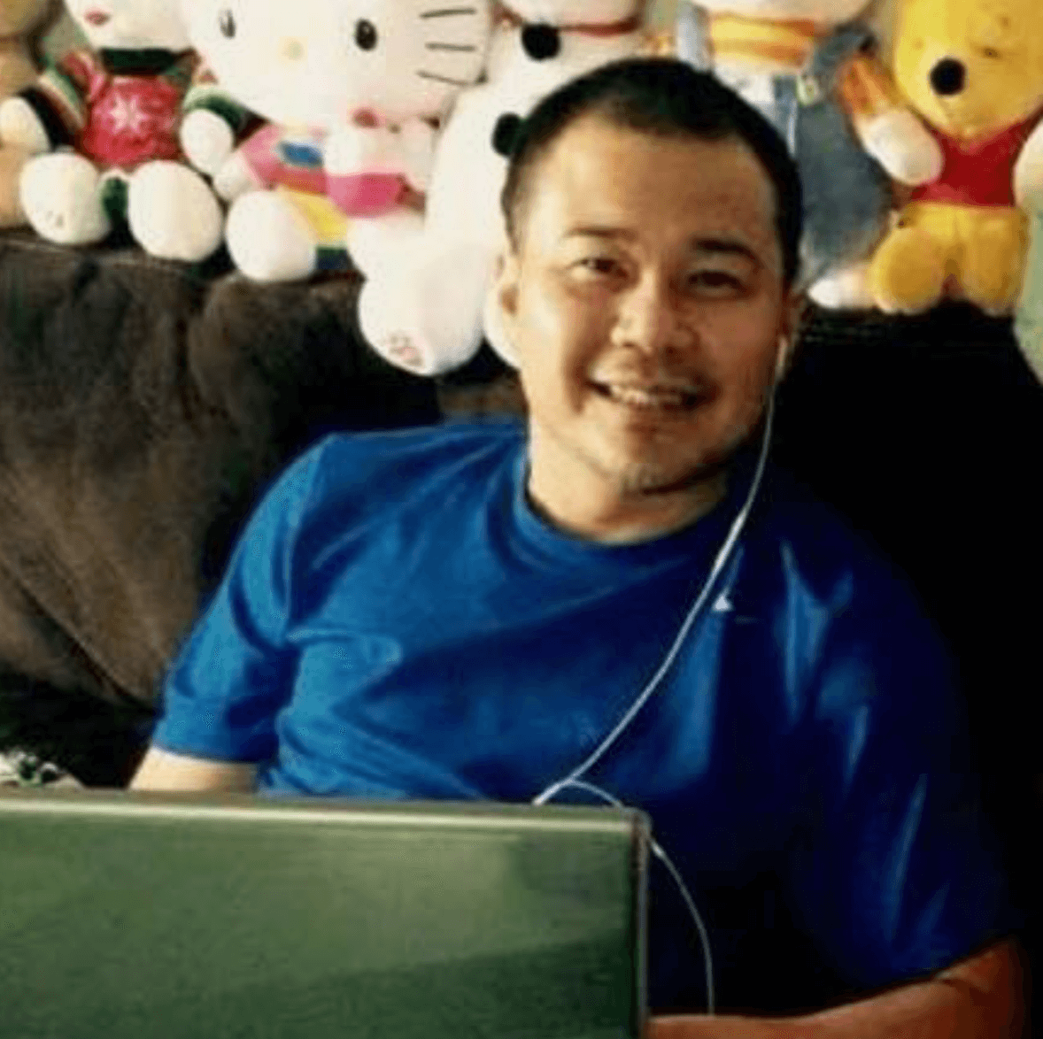 Man in a blue shirt smiling on a couch, with stuffed animals in the background