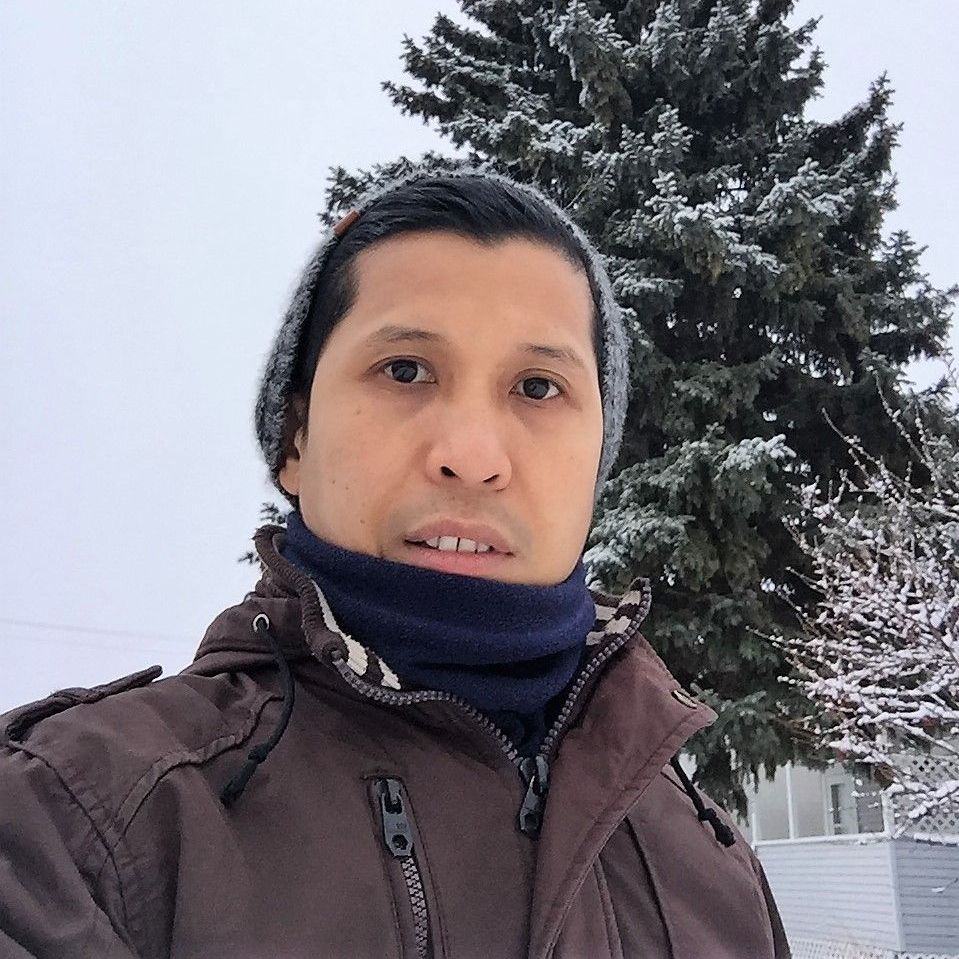 Person in a winter jacket and beanie taking a selfie outdoors in the snow beside a pine tree.