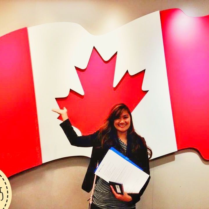 Person smiling and pointing at a large red maple leaf on a Canadian flag backdrop, holding papers