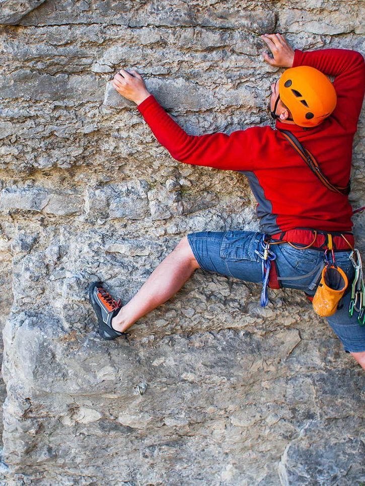 Rock climber in red shirt and orange helmet scaling a gray cliff face.
