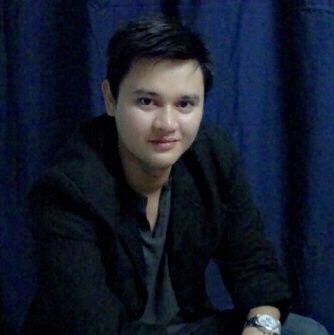 Person in a black jacket seated against a dark blue curtain, looking at the camera with a slight smile.