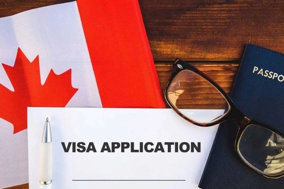 Visa application form with Canadian flag, passport, eyeglasses, and pen on a wooden desk.