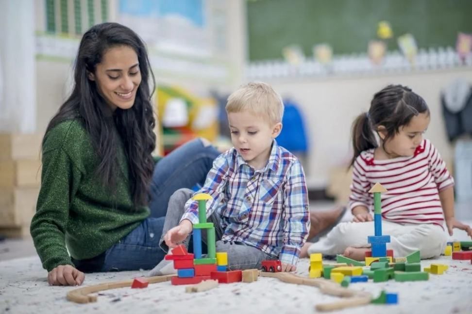 Adult and two children playing with colorful blocks on the floor in a bright room