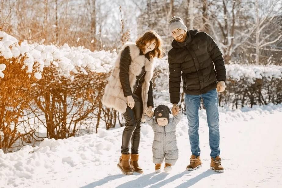 Family standing together in a snowy park, bundled in winter coats beside a hedge and bare trees.