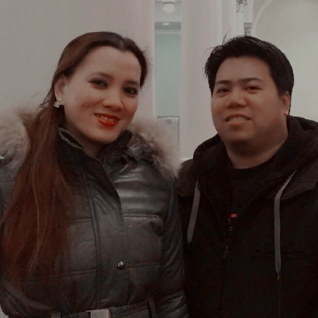 Two people smiling indoors, wearing dark winter jackets, standing close together.
