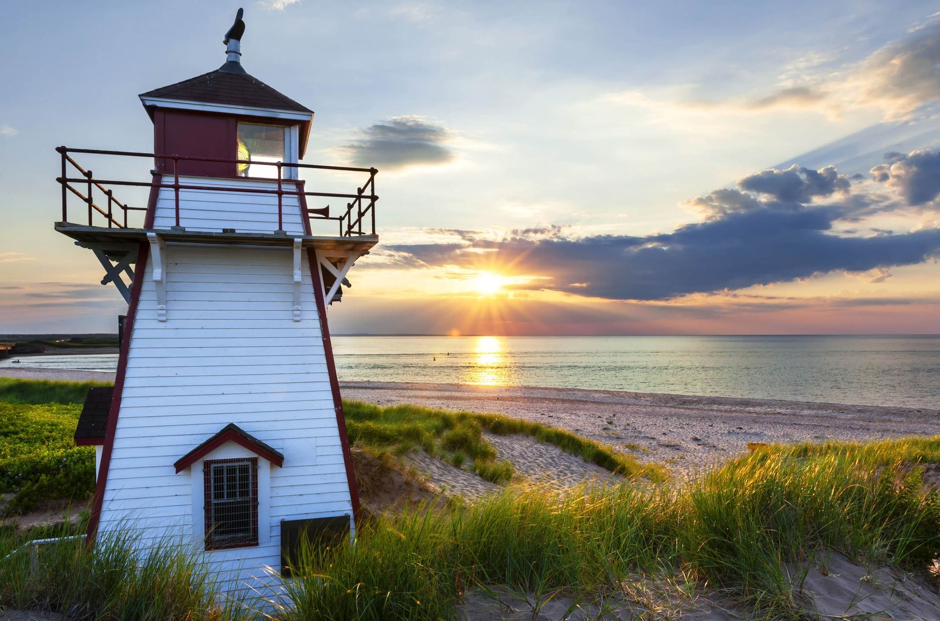 White lighthouse at sunset by a grassy shoreline with calm water and dramatic clouds.