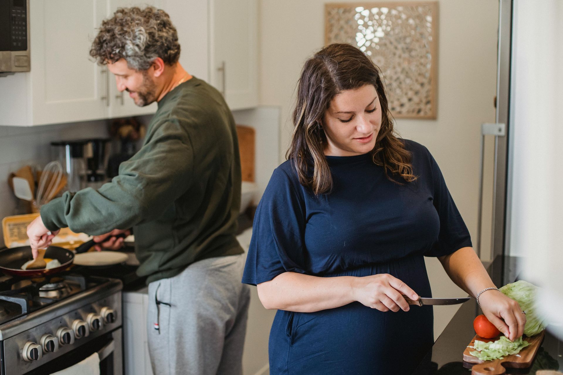 Two people preparing food in a kitchen, one cooking at the stove and one slicing vegetables.