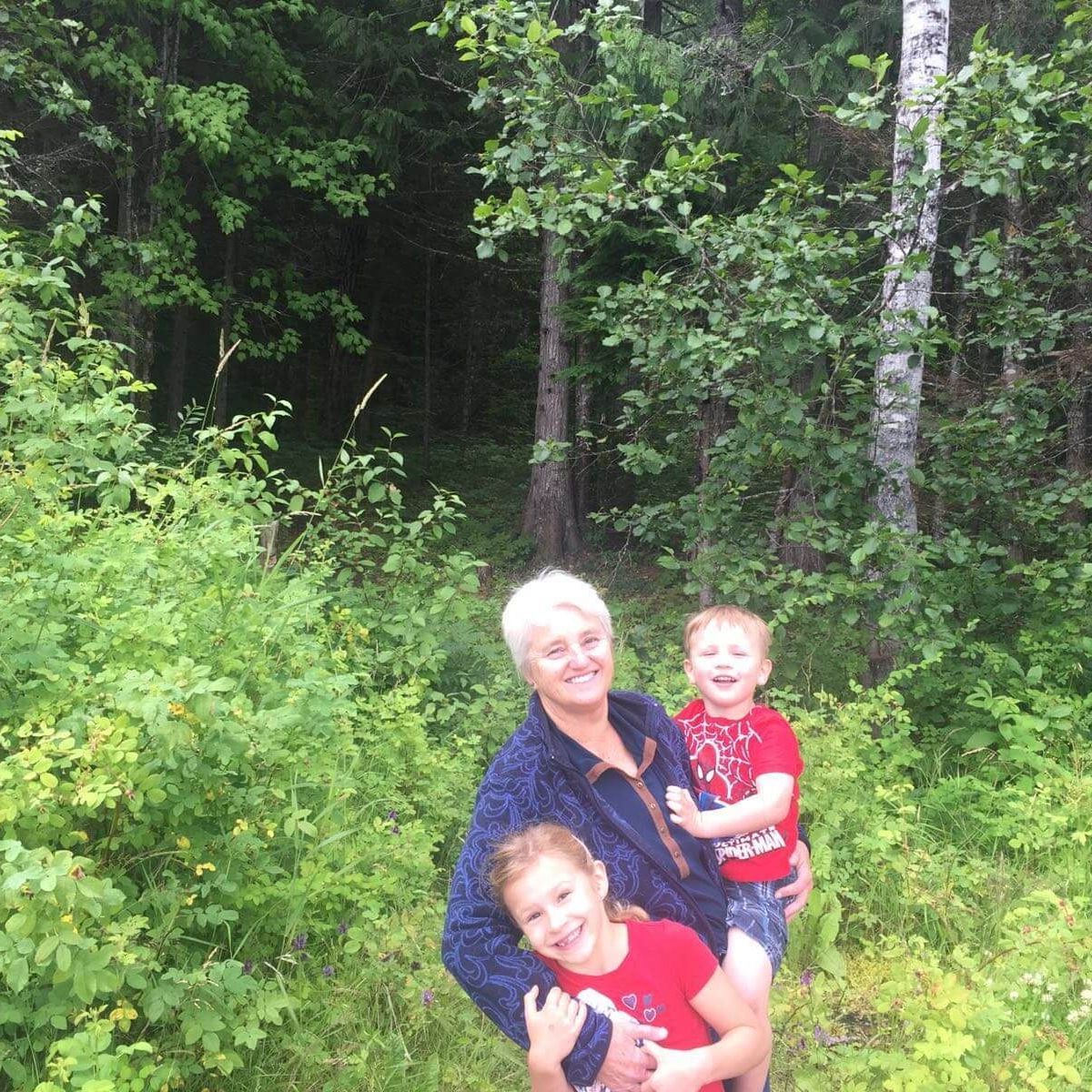 Two children and a woman smiling in a leafy forest clearing, one child in red in the foreground