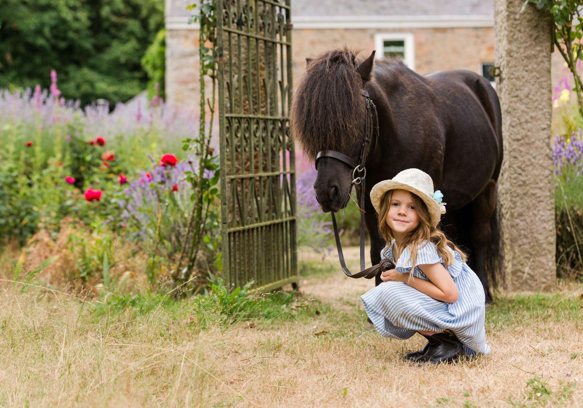 Girl in a sun hat crouches next to a black pony, near a garden gate.