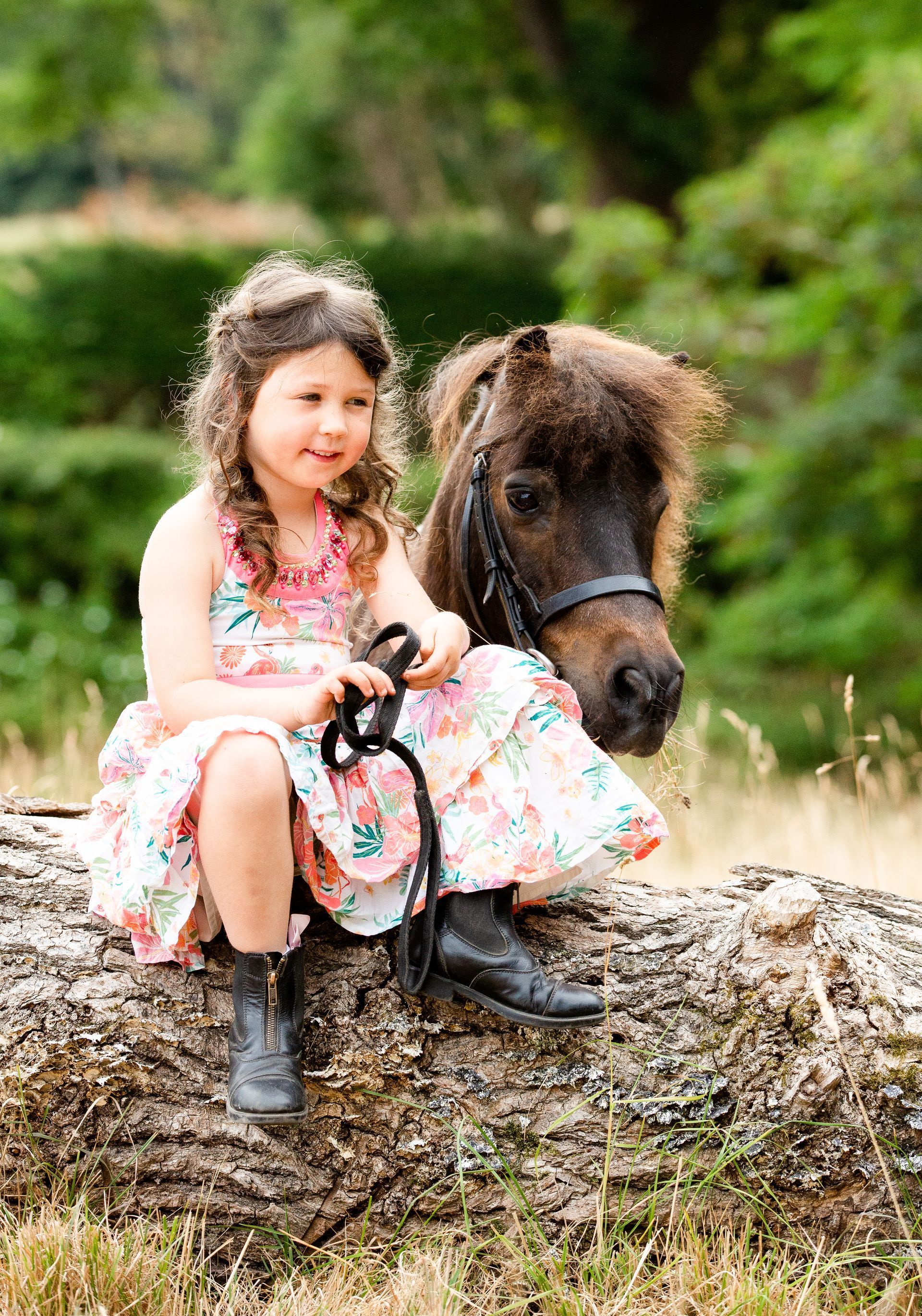 Girl in floral dress sits on a log with a miniature horse, smiling.
