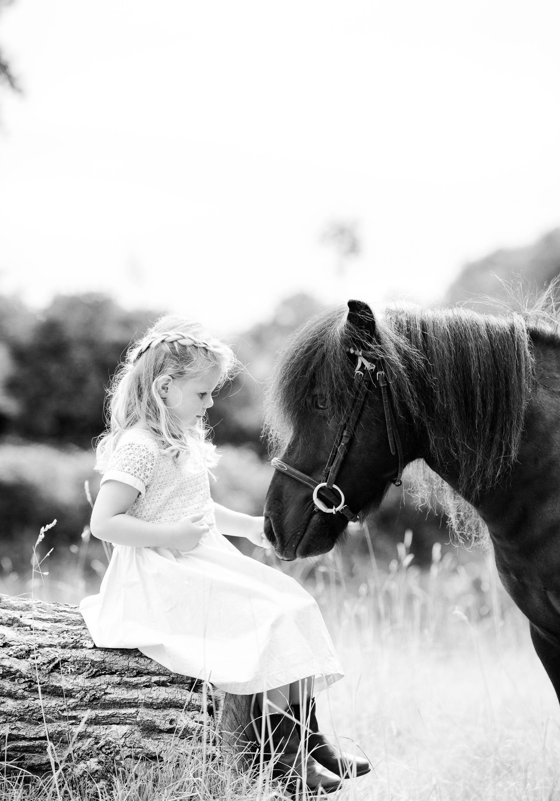 Girl in white dress sitting on log, petting pony with a dark mane, outdoors.