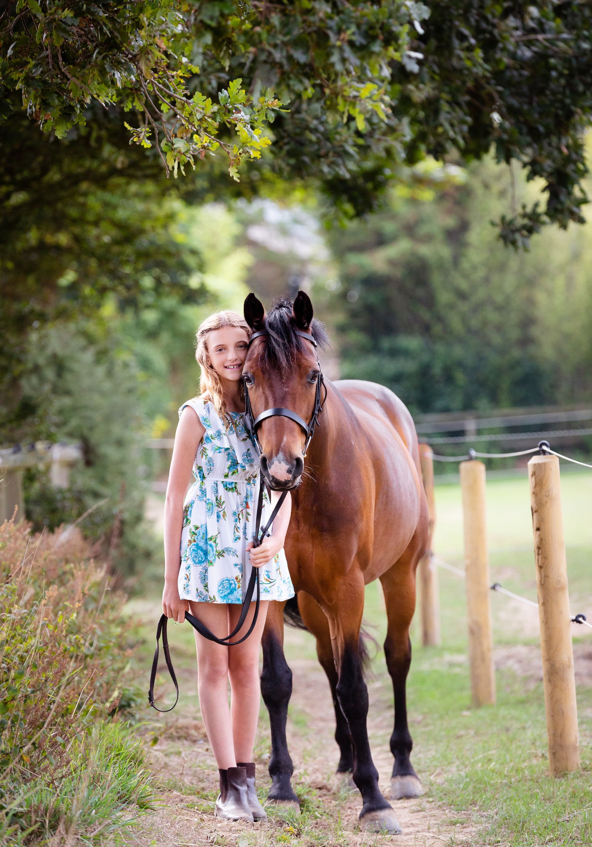 Woman in floral dress standing beside a brown horse on a path, trees in the background.