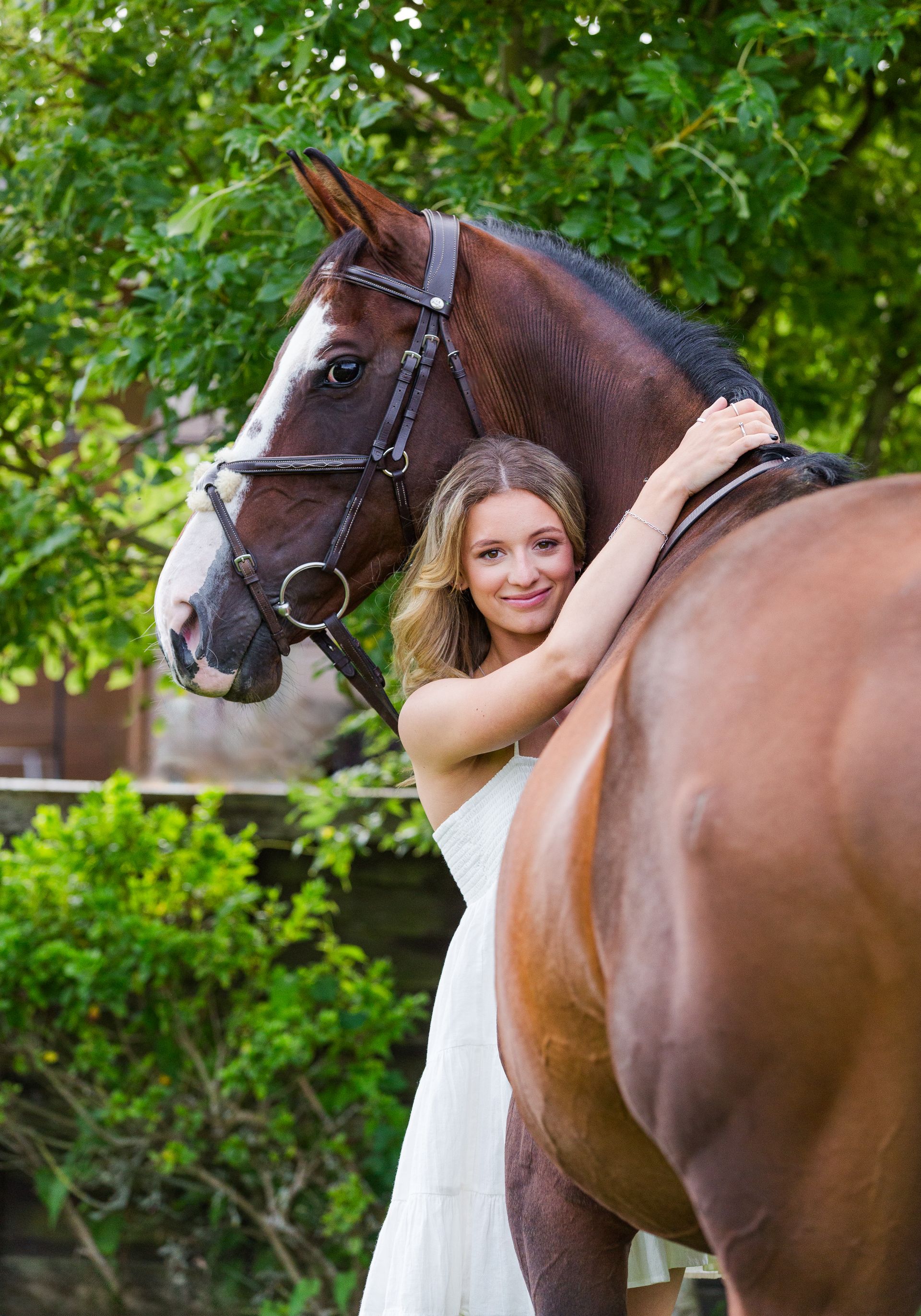 Woman in white dress embraces a brown horse outdoors near green foliage.
