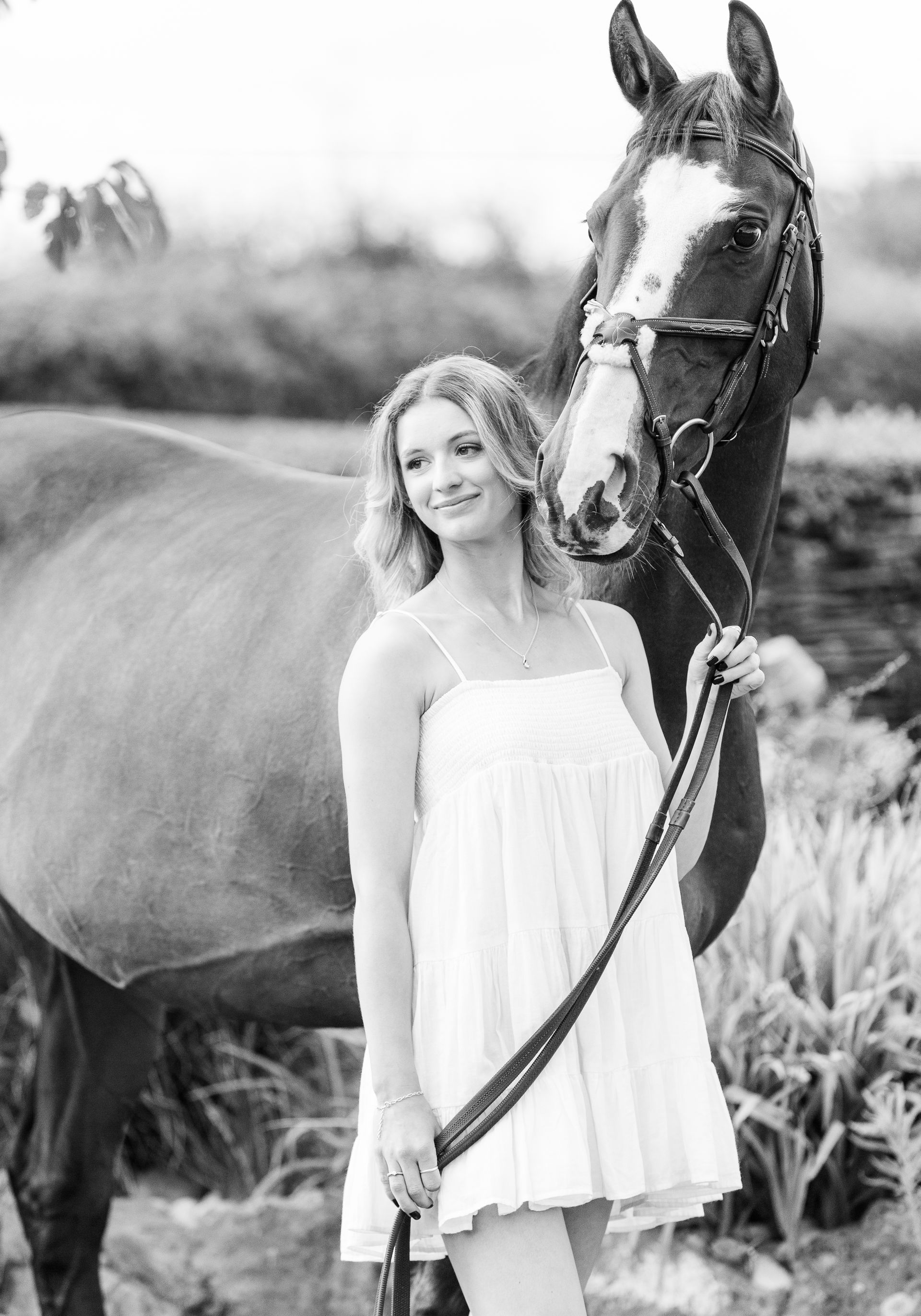 Woman in white dress smiles, holding a horse's reins. Horse has bridle. Outdoor setting. Black and white.