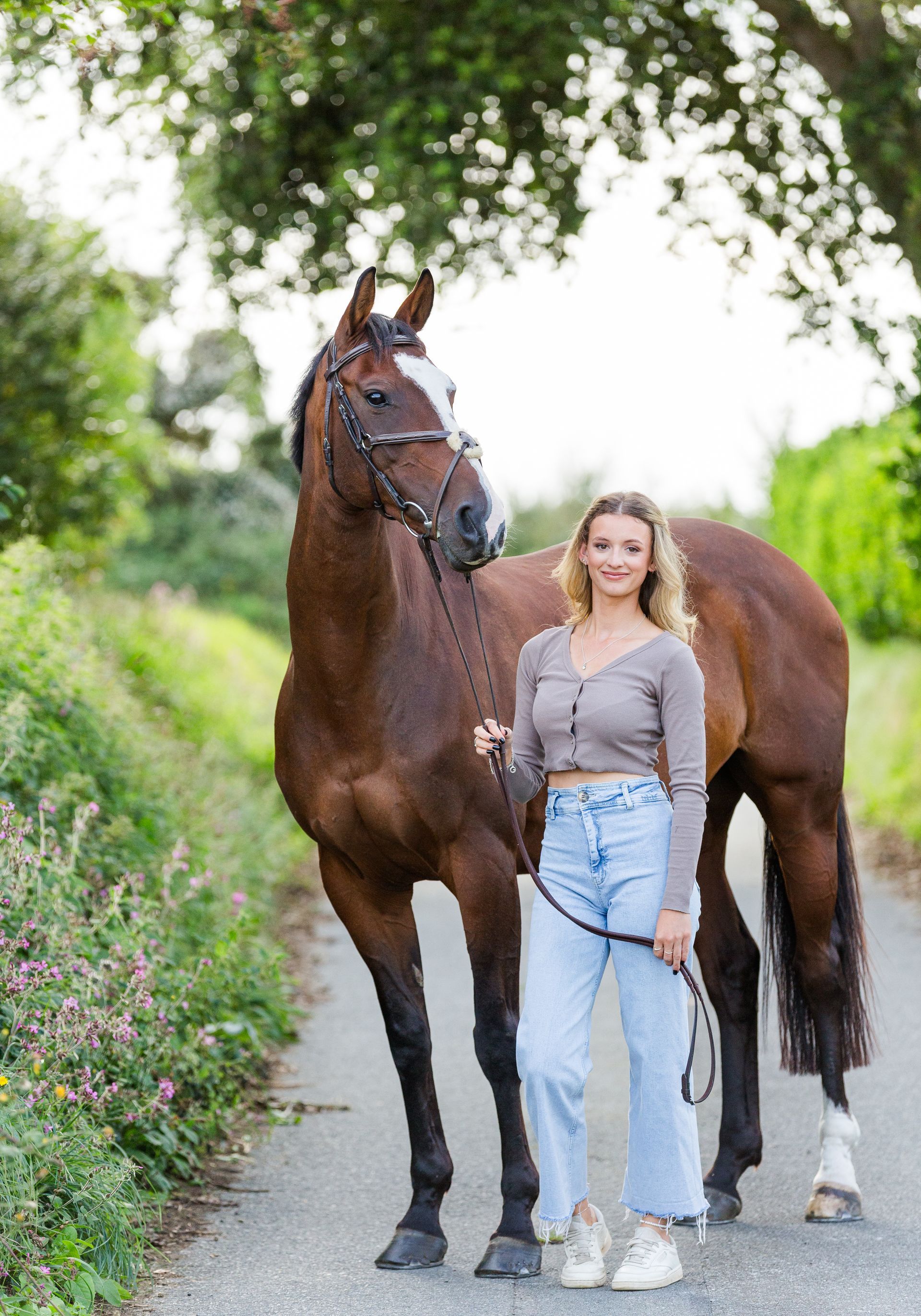 Woman standing with a brown horse on a path. She holds the reins. Overcast sky and greenery in the background.