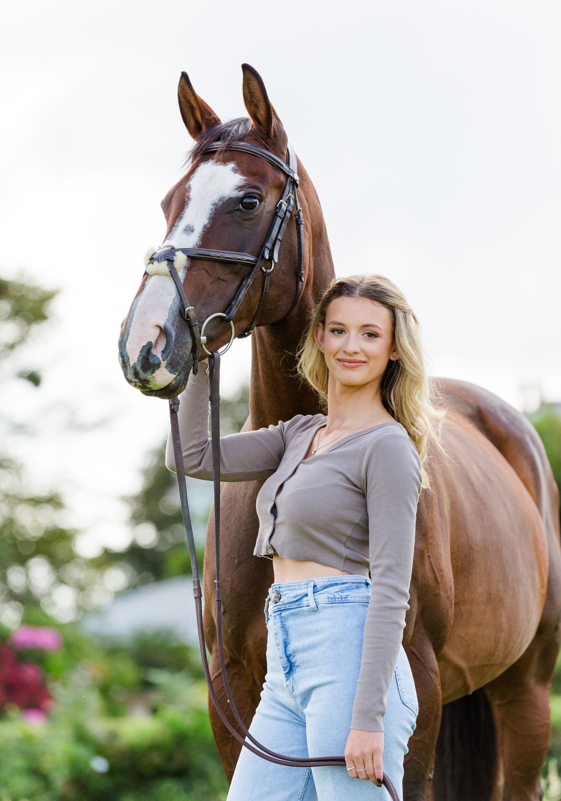 Woman in jeans and cropped sweater stands with a brown horse in a field.