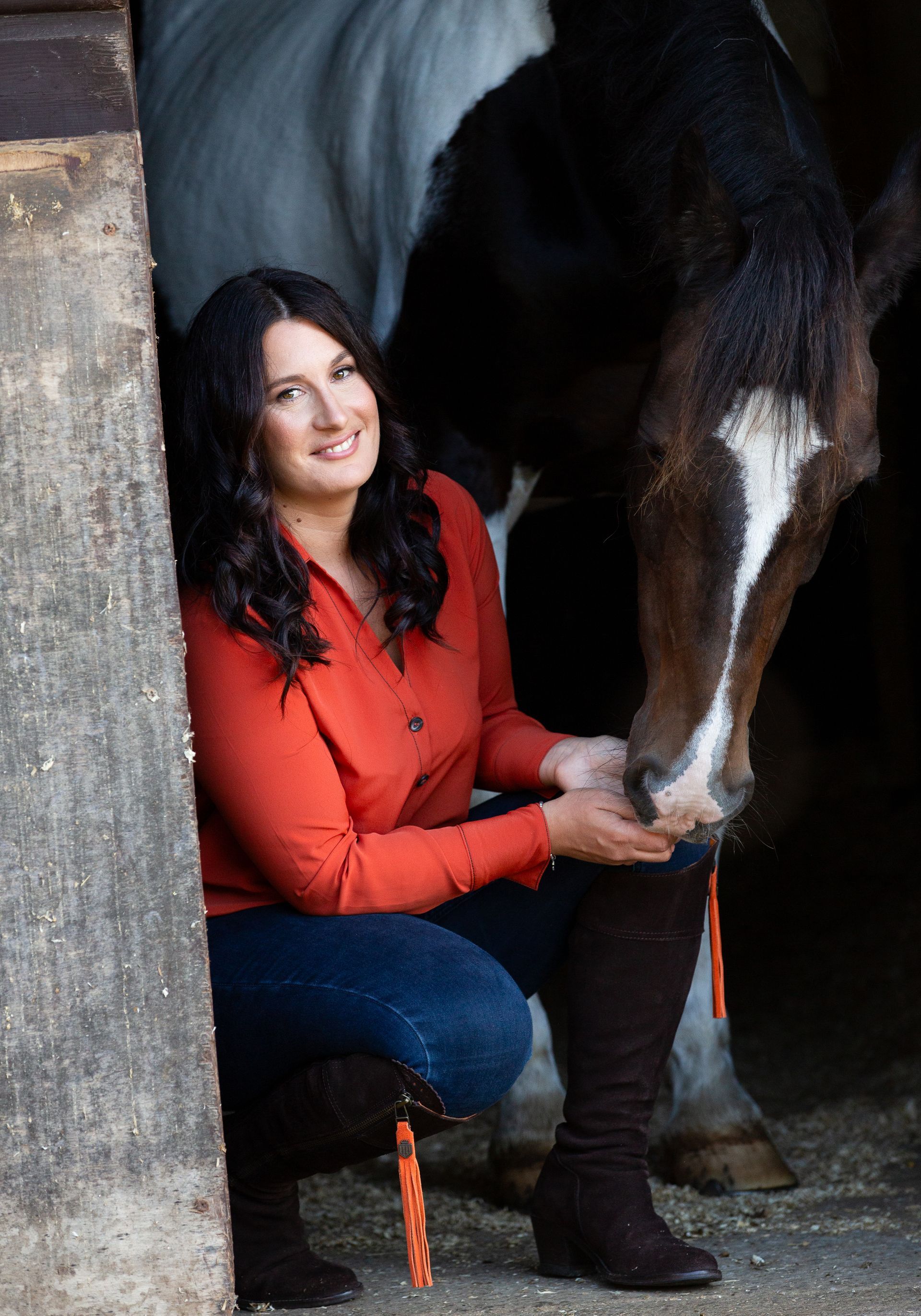 Woman kneeling, petting a horse's head in a barn, wearing an orange shirt and jeans.