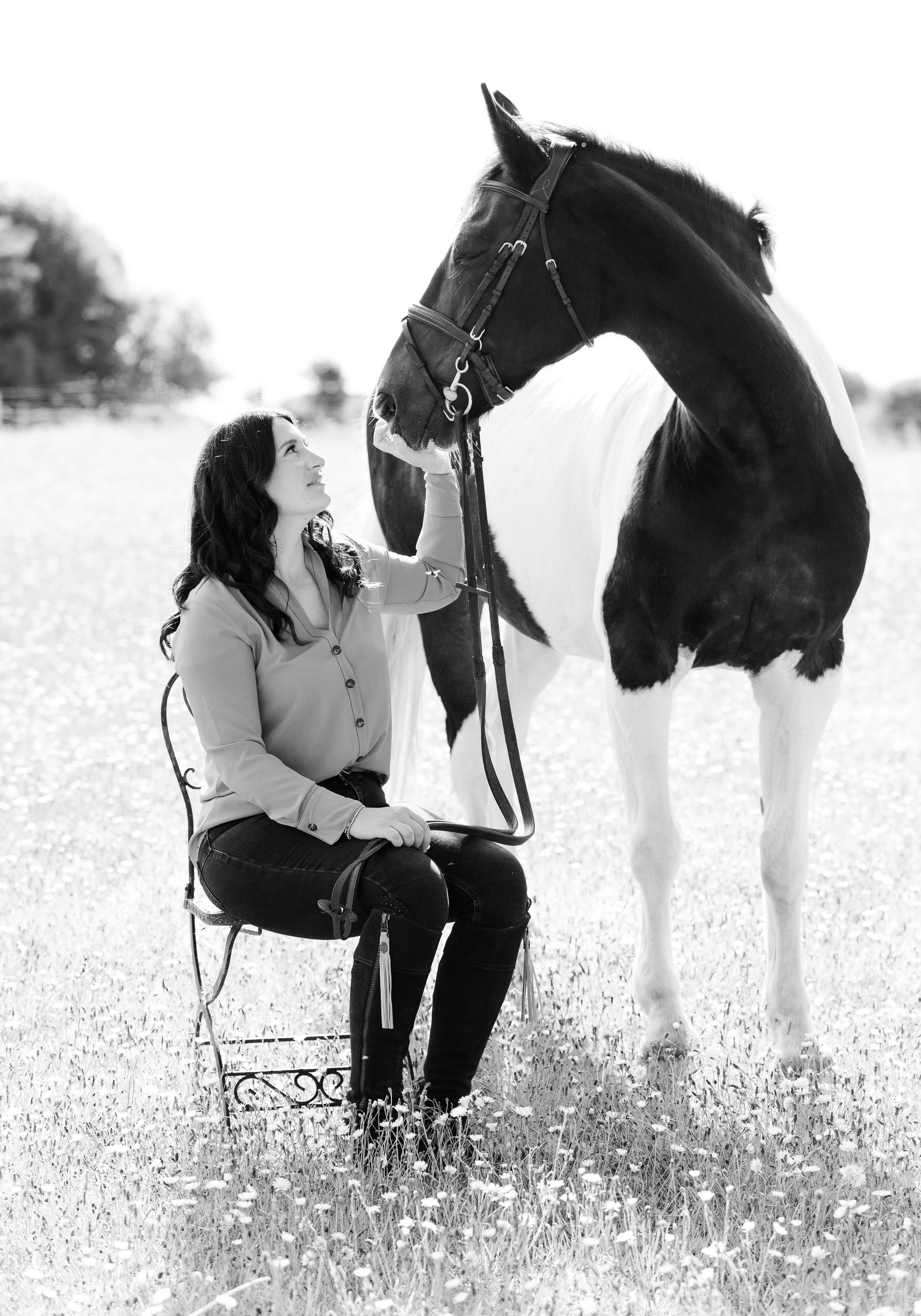 Woman seated, petting a black and white horse in a field of flowers.