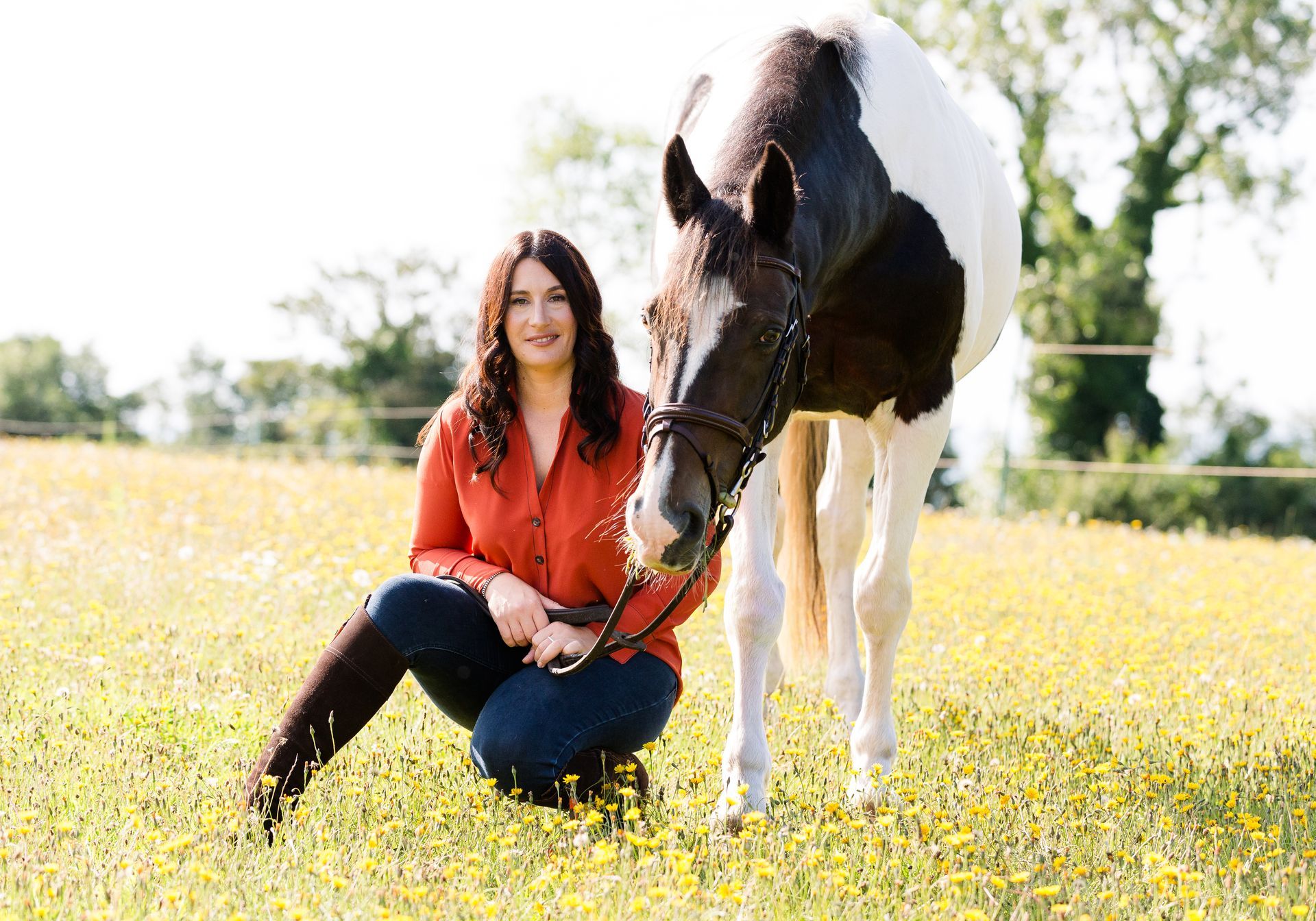 Woman in orange shirt kneels next to a piebald horse in a sunny field of yellow flowers.