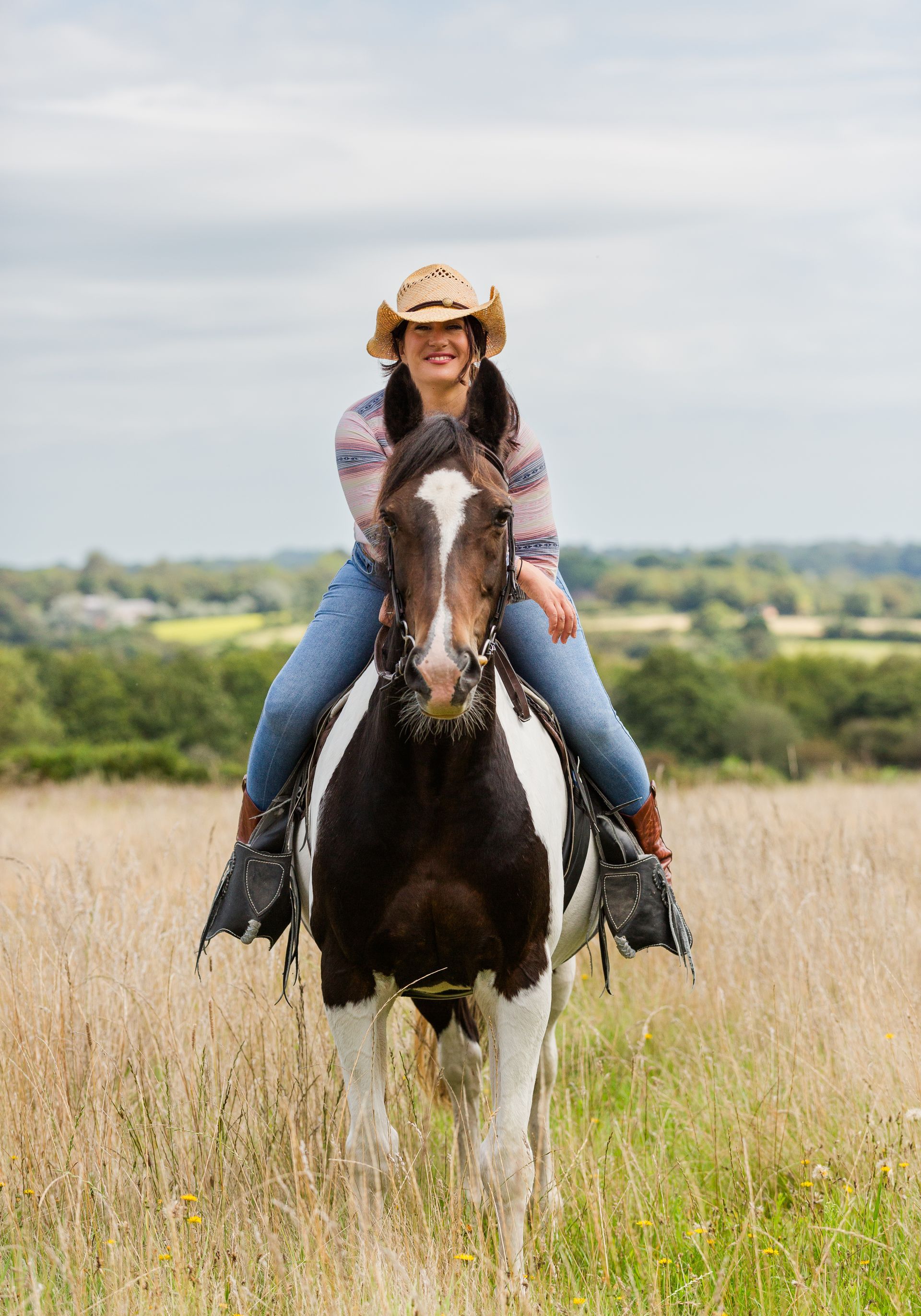 Woman wearing a cowboy hat rides a black and white horse in a field, smiling.