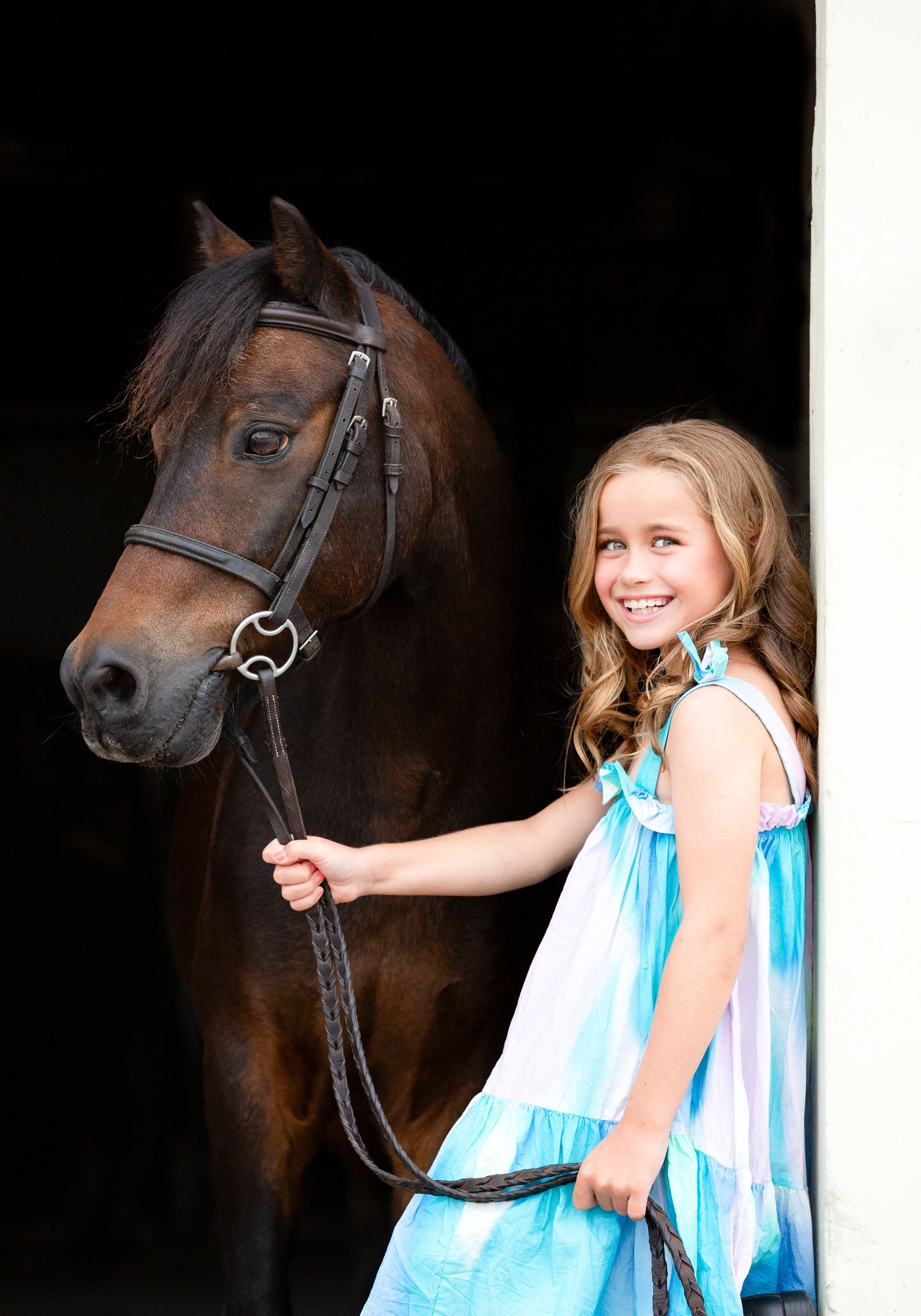 Girl with brown pony, smiling, holding reins. They stand in a dark doorway, girl wearing a blue and white dress.